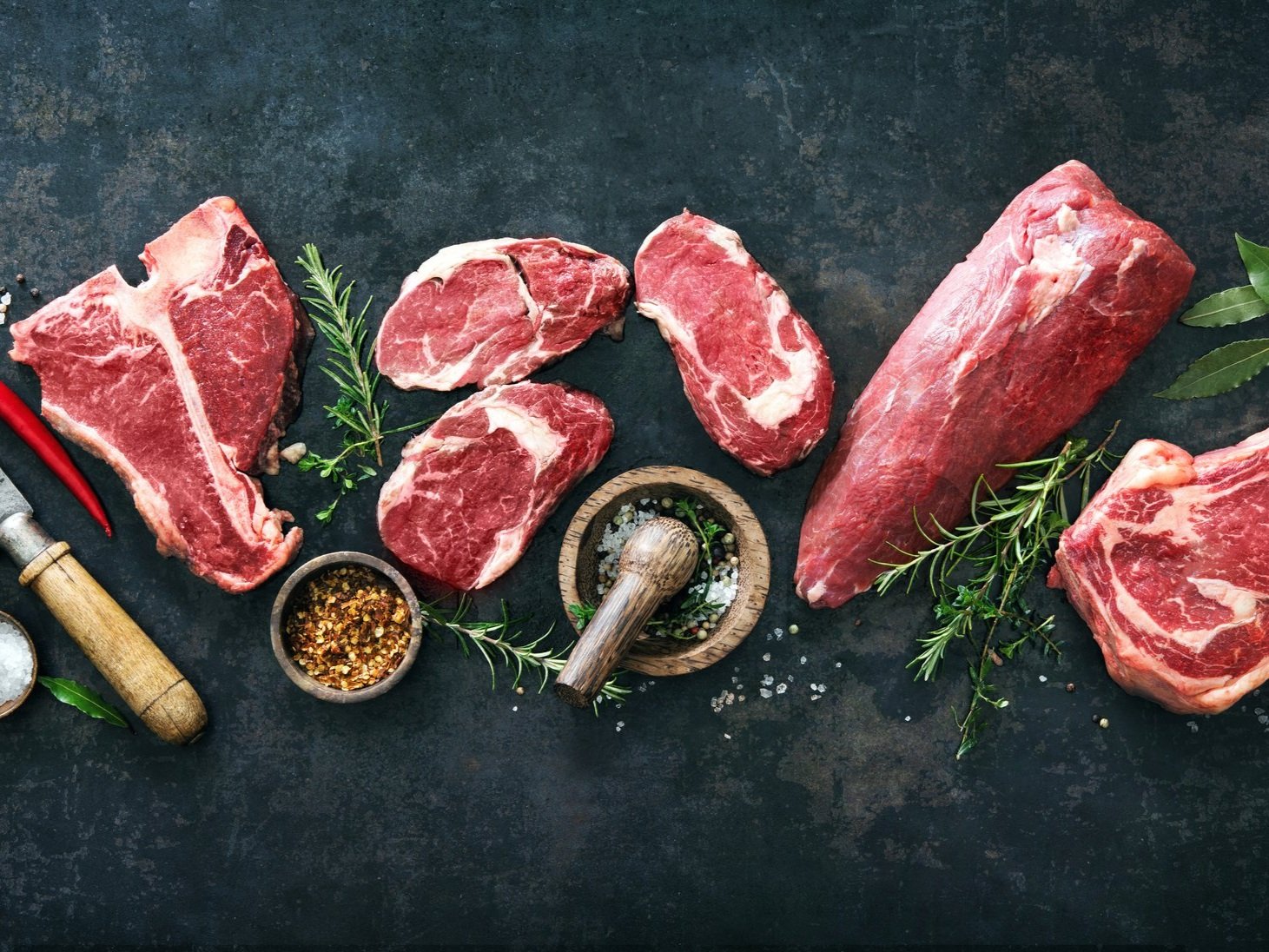 Assorted cuts of fresh red meat arranged on a dark surface, accompanied by herbs, spices, and a wooden mortar and pestle, ideal for culinary presentations and recipes.