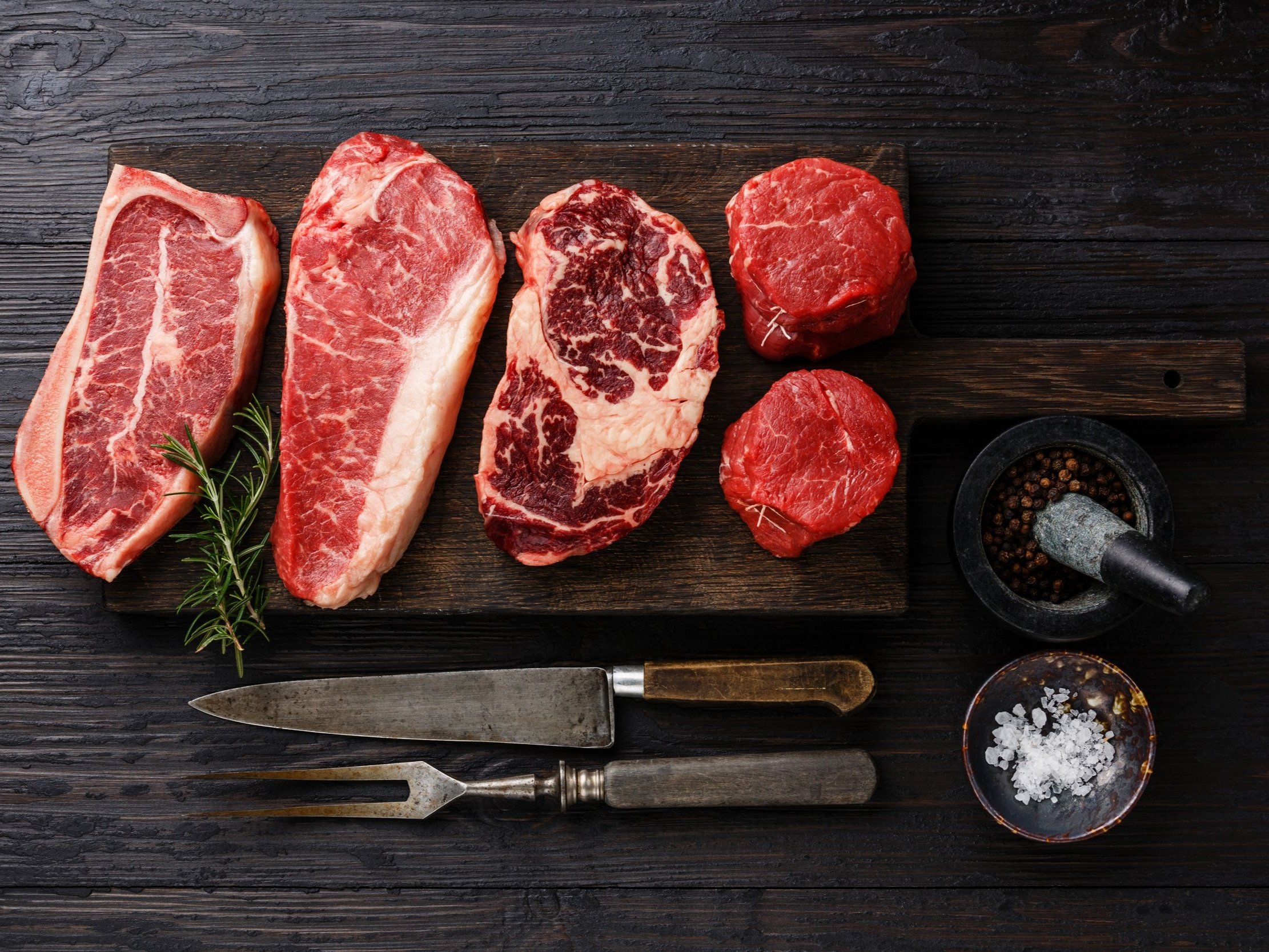Assorted cuts of raw beef, including ribeye, sirloin, and tenderloin, arranged on a wooden cutting board with fresh rosemary, a mortar and pestle with peppercorns, and a small bowl of sea salt, highlighting gourmet cooking ingredients.
