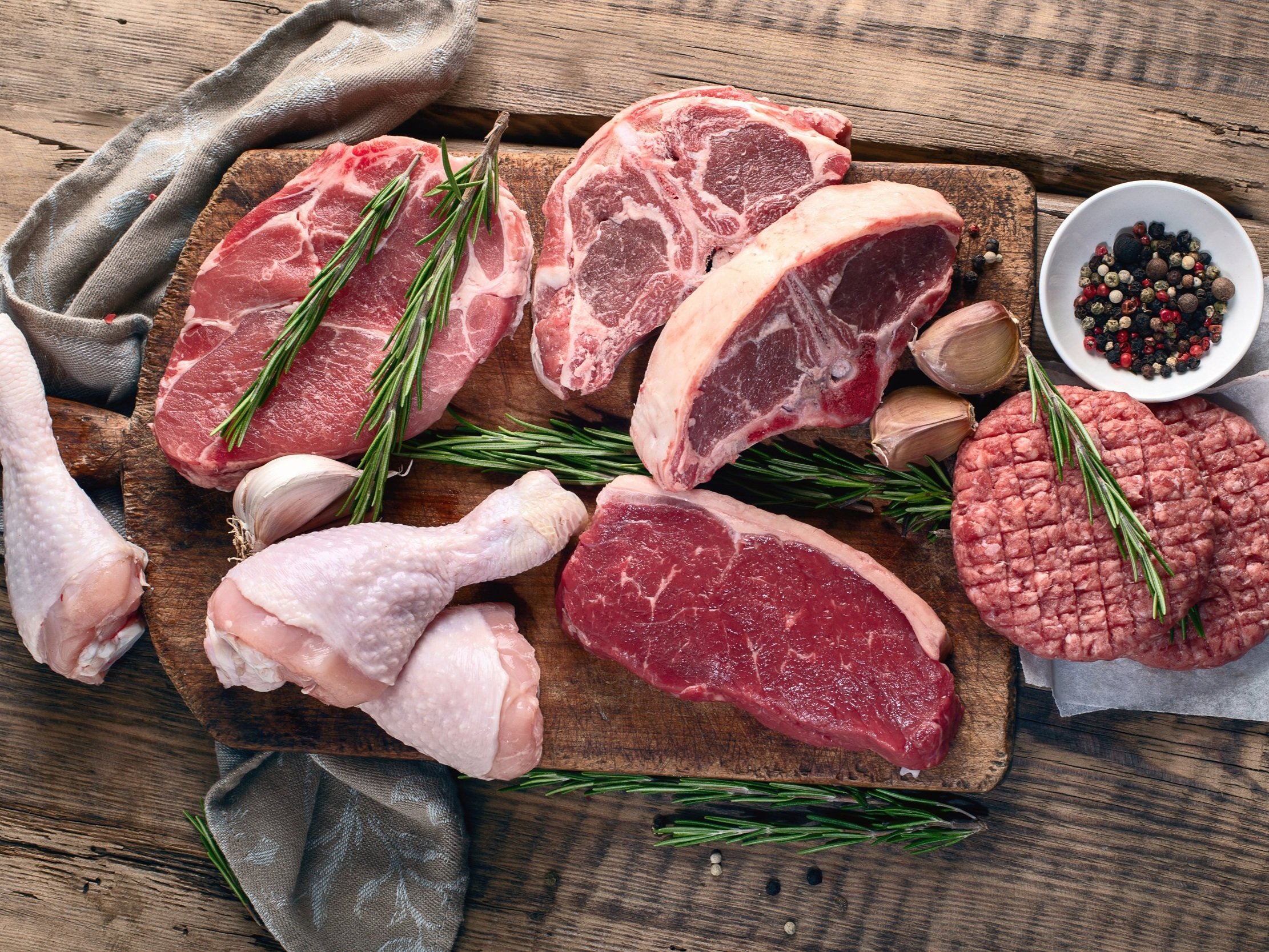 Assorted raw meats including chicken drumsticks, beef steaks, pork chops, and ground beef on a wooden cutting board, garnished with fresh rosemary and surrounded by garlic and a bowl of mixed peppercorns.