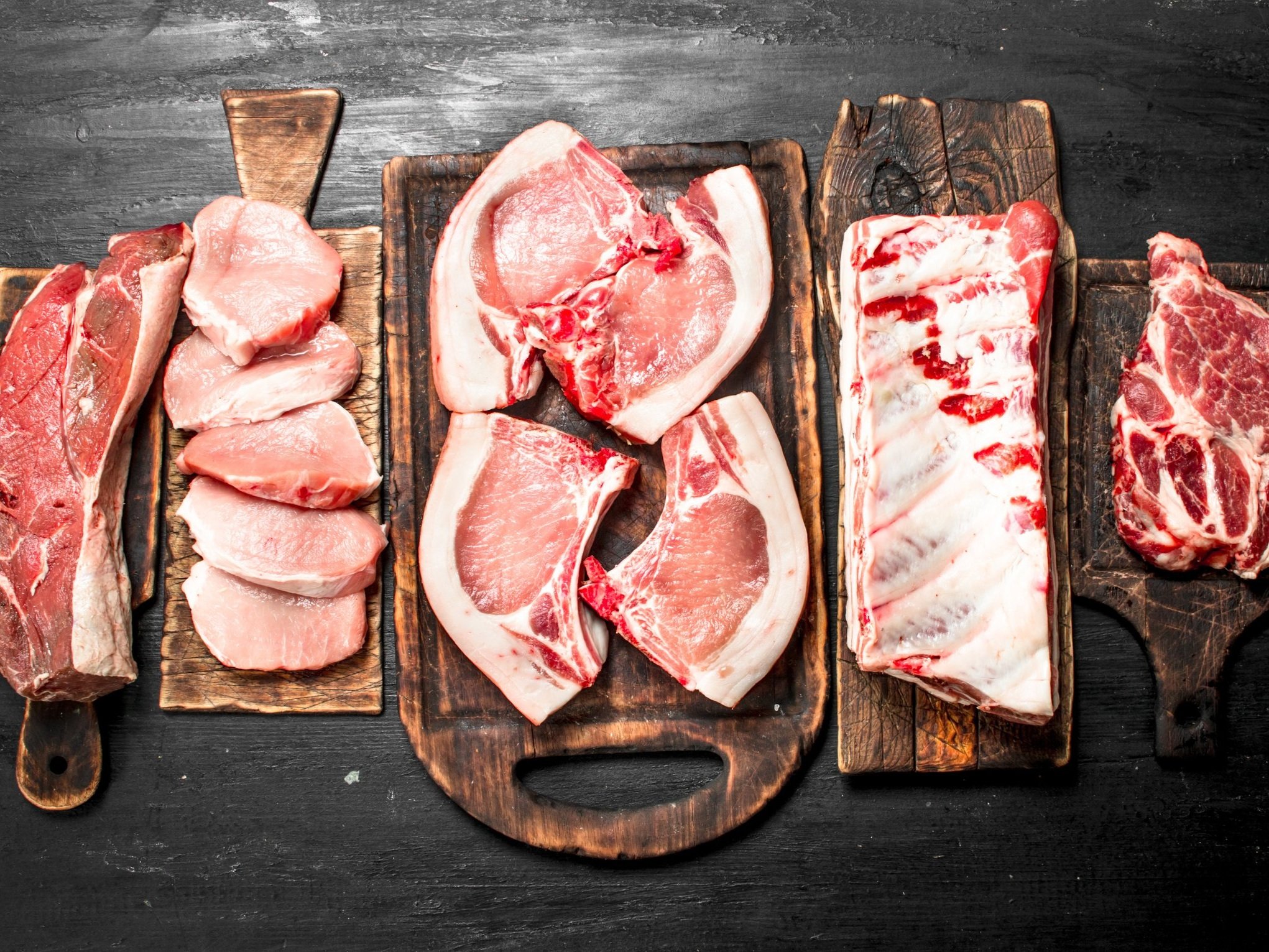 Assorted cuts of fresh meat displayed on wooden cutting boards, including pork chops, ribs, and steaks, arranged on a dark surface for a culinary presentation.